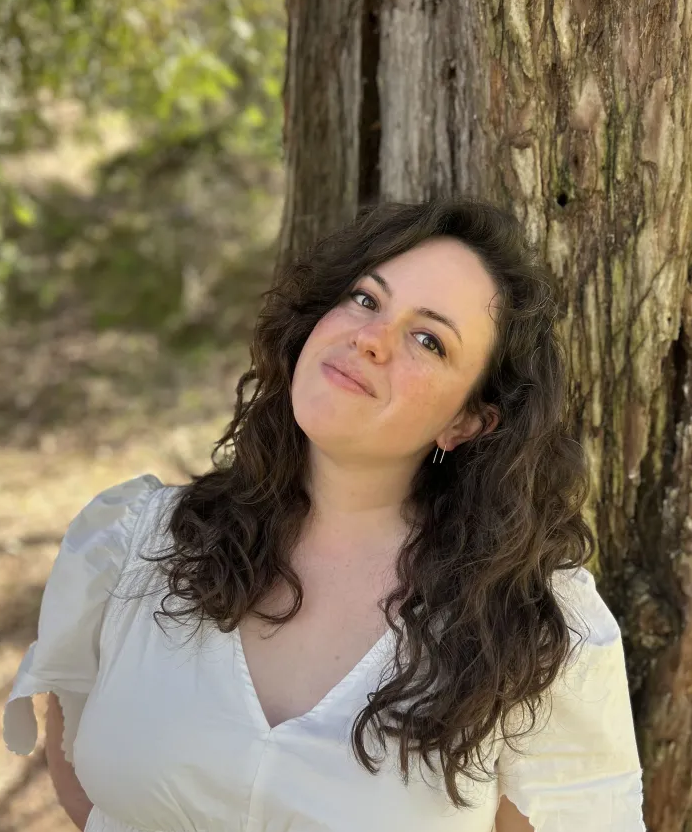 Rachel Wilman stands against a tree with long curly hair in a white dress